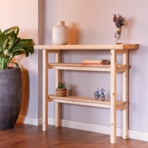 Consola de recibidor pieza única Whare tapu de madera clara con dos estantes, decorada con libros y una planta al lado