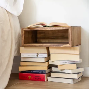 Mesita de noche flotante de madera Pa en tono oscuro sobre pilas de libros junto a la cama