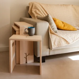 Set de mesas apilables de madera Tāpae junto a un sofá con manta y cojín amarillo, con una taza encima de la mesa más pequeña